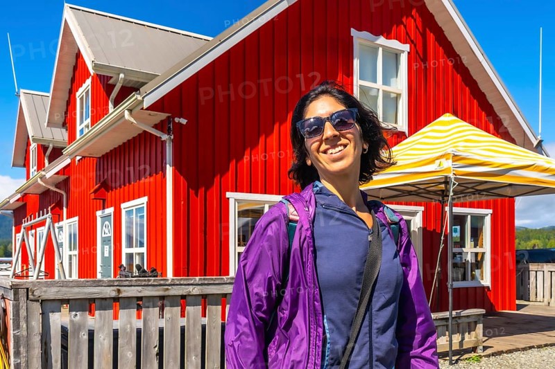 Happy tourist wearing sunglasses and purple jacket smiling in front of a vibrant red building in tofino, a popular tourist destination on vancouver island, british columbia, canada, during summer