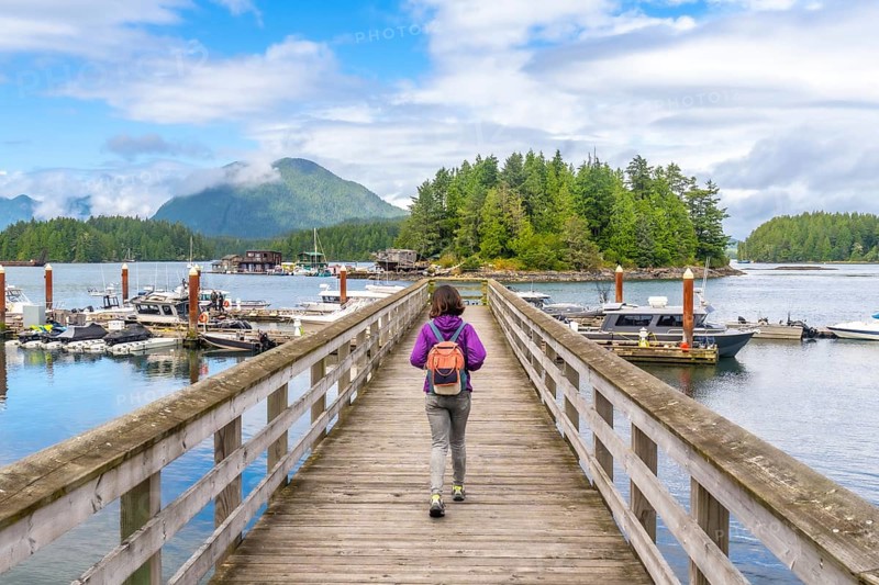 Female tourist with backpack walking down a wooden pier in tofino, british columbia, with boats, harbor, and forested island in background on a sunny summer day