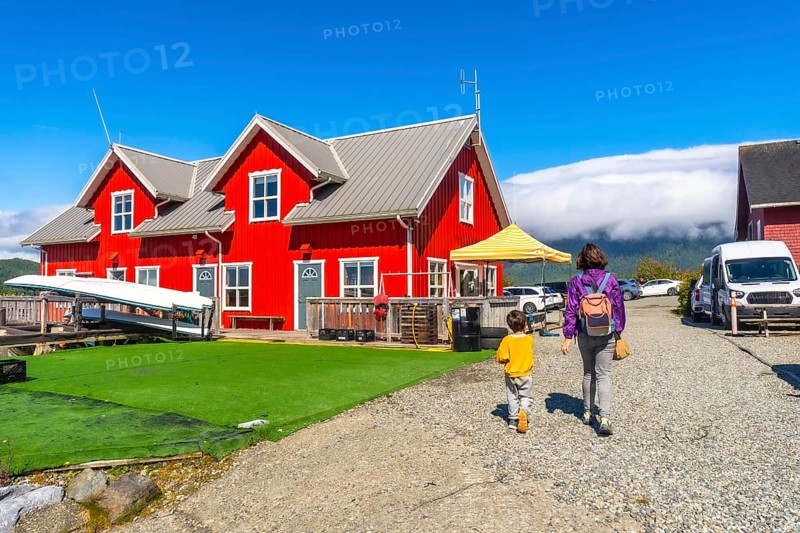 Mother and son are walking on a gravel path near a vibrant red house with white trim, canoes resting on a green turf under a clear blue sky in tofino, vancouver island