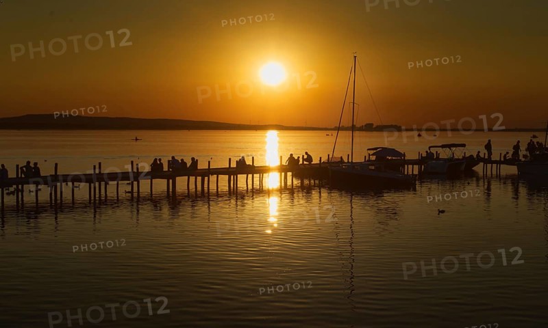 Sunset over the Steinhuder Meer with people on a pier, reflecting sailboats, peaceful, Steinhude
