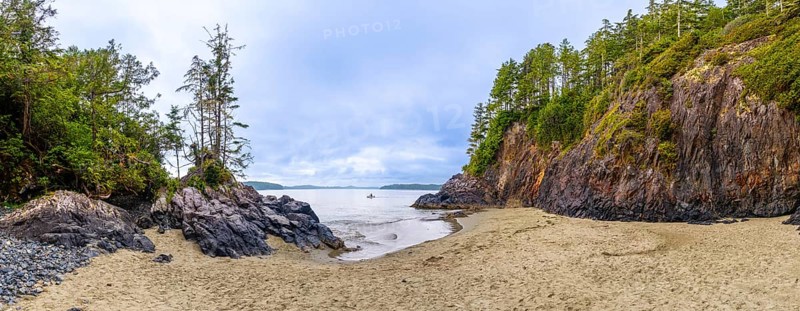 Panoramic view of a secluded beach and inlet in tofino, vancouver island, featuring dramatic rock formations, lush green vegetation, and a lone kayaker enjoying the serene waters under a cloudy sky