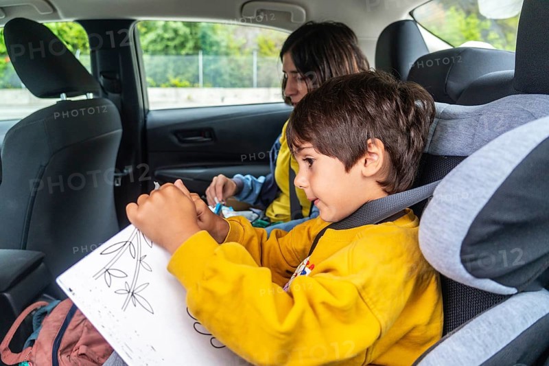 Child concentrating on drawing while sitting in a car booster seat, his mother preparing medicine in the background, creating a tender moment of childhood amidst travel