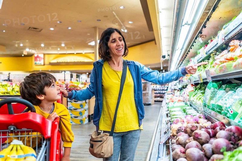 Mother points to fresh vegetables while shopping with her son in the produce section of a grocery store, promoting healthy eating habits and family bonding