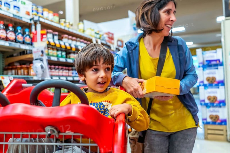 Mother holding a box of cookies next to her son sitting in a shopping cart, enjoying their grocery shopping experience together in a brightly lit supermarket