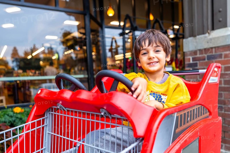 Smiling preschooler having fun sitting in a special children's shopping cart shaped like a firetruck, enjoying a shopping trip with family outside a grocery store