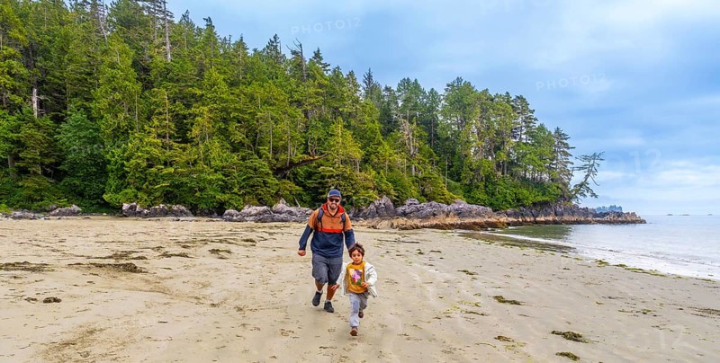 Father and son running together on a sandy beach with lush green forest and rocky coastline in tofino, vancouver island, british columbia, canada, enjoying quality family time in nature