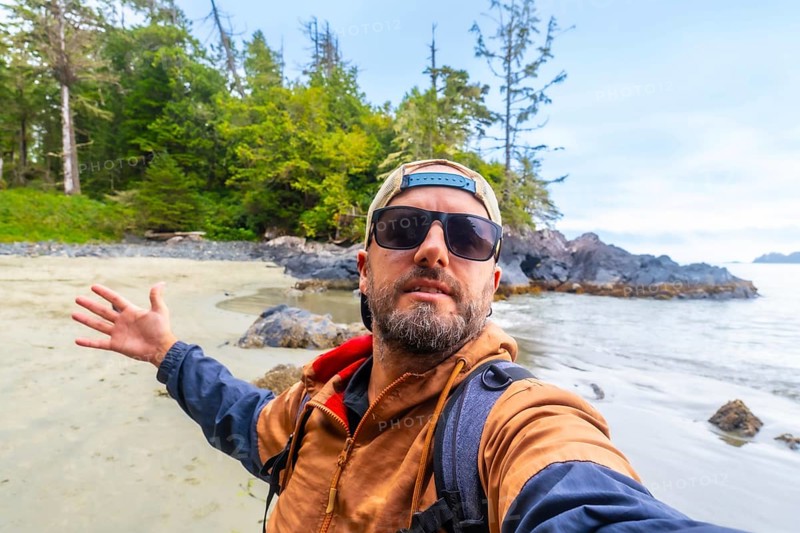 Tourist with sunglasses and baseball cap taking a selfie on chesterman beach, showing the beautiful landscape of tofino on vancouver island, british columbia, canada, during a sunny day