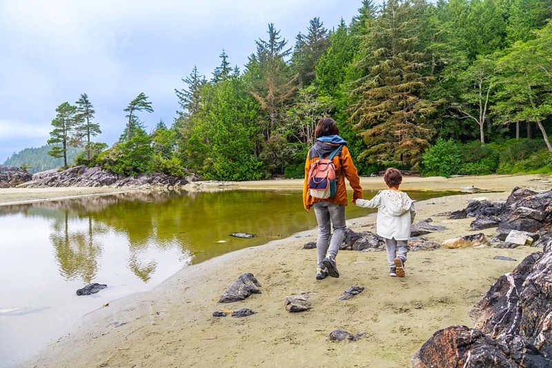 Mother and child enjoying a leisurely stroll along a sandy beach in tofino, british columbia, surrounded by lush forests and calm reflective waters