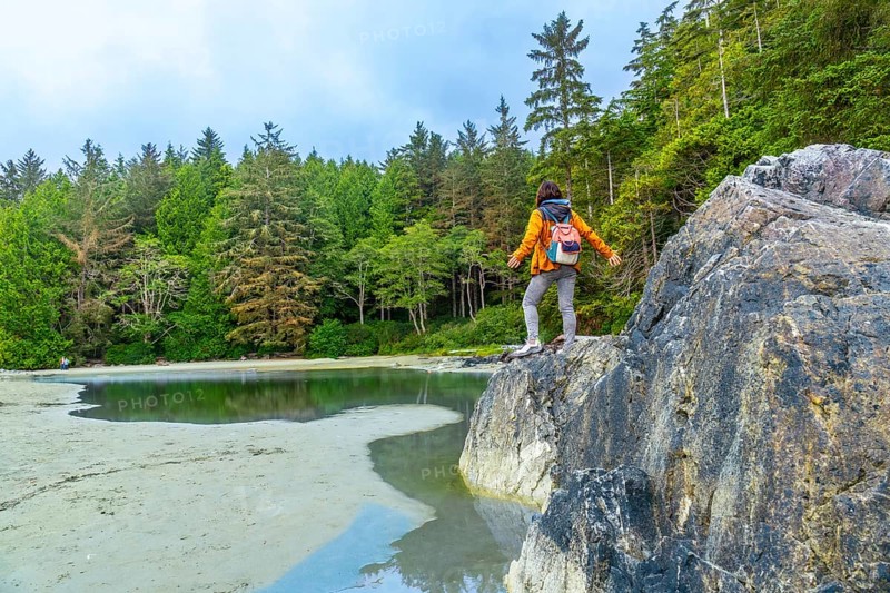 Female hiker with a backpack standing on rocks with outstretched arms, savoring the breathtaking view of mackenzie beach and the surrounding lush forest on vancouver island
