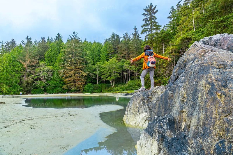 Tourist with open arms is standing on a rock enjoying the beautiful scenery of a beach in tofino. A district municipality in british columbia. Canada. Located on the west coast of vancouver island