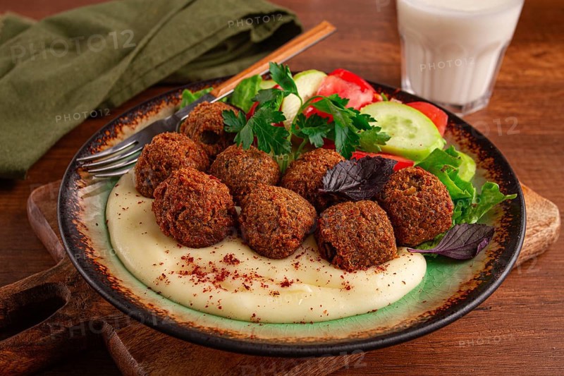 A plate holds falafel balls, tahini, salad with tomatoes and cucumbers, and a drink. The setting is warm and inviting, perfect for a midday meal with fresh ingredients