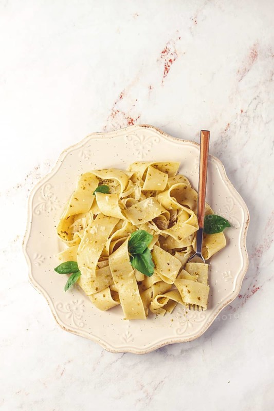Fresh papardelle pasta, with basil and pesto sauce, is served, on a white plate, with a fork held by hand