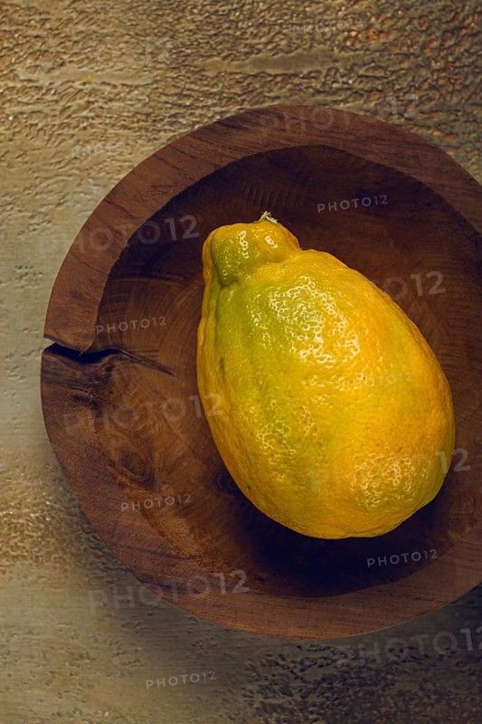 A vibrant yellow fruit, possibly a lemon or a ripe papaya, sits in a handcrafted wooden bowl against a textured background. Natural light highlights its glossy skin and freshness