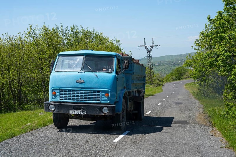 A blue truck drives on a sunny country road surrounded by trees and a power pole, MAZ truck, exhaust gases, Armenia