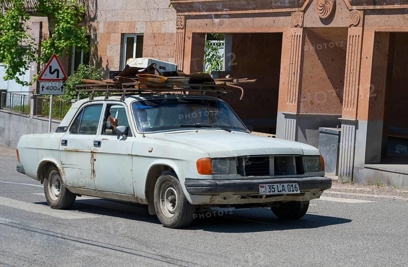A white car with roof luggage drives through an urban environment with old buildings, GAZ Volga, Armenia