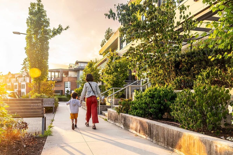 Mother and son holding hands are walking away from the camera on a sidewalk in a residential neighborhood of vancouver, with warm sunset light illuminating the scene