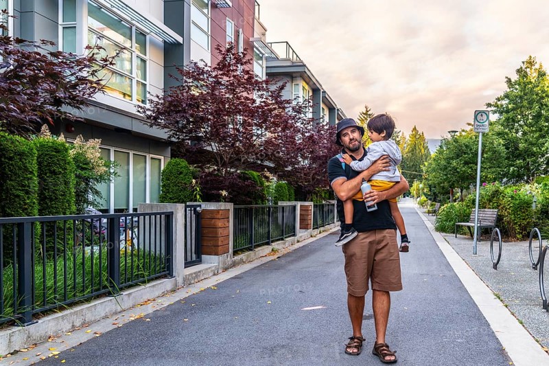 Father carrying his son while walking on a sidewalk in a residential area of vancouver, british columbia, with modern townhouses, trees, and a bus stop sign in the background during a sunny day