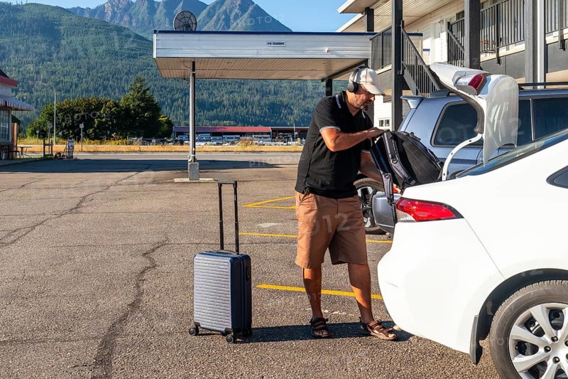 Tourist wearing headphones is loading luggage into car trunk in motel parking lot, preparing for road trip adventure in scenic mountain landscape
