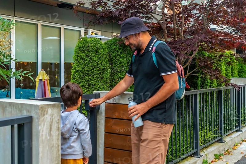 Father holding a reusable water bottle and wearing a bucket hat opens the gate for his son at a daycare in vancouver, british columbia, starting their morning drop off routine