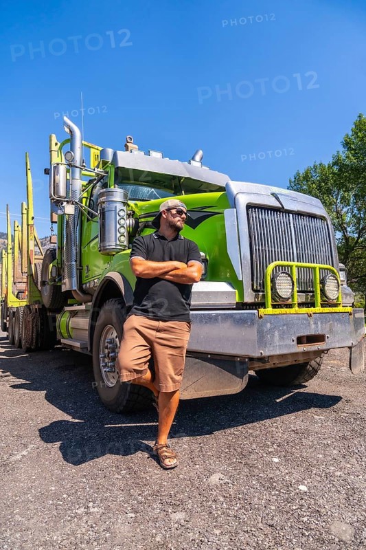 Truck driver confidently posing with arms crossed, leaning against a modern green logging truck in kamloops, british columbia, under a bright sunny sky on a warm summer day