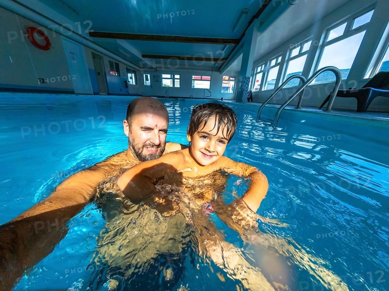 Father and son enjoying a playful moment while taking a selfie in an indoor swimming pool, creating lasting memories during their leisure time together