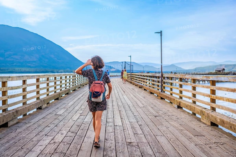 Young woman with backpack walking down a long wooden pier, enjoying the scenic view of the shuswap lake and surrounding mountains in salmon arm, british columbia, canada
