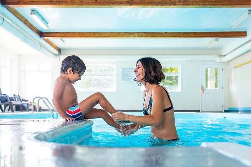 Happy mother and son enjoying playful moments at the edge of an indoor swimming pool, splashing water and sharing laughter during a fun filled leisure activity, creating joyful memories together
