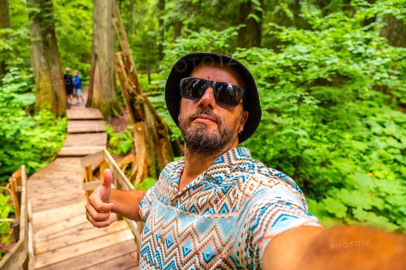Happy tourist wearing sunglasses and bucket hat taking a selfie while hiking the beautiful giant cedars boardwalk trail in mount revelstoke national park, british columbia, canada, giving thumbs up