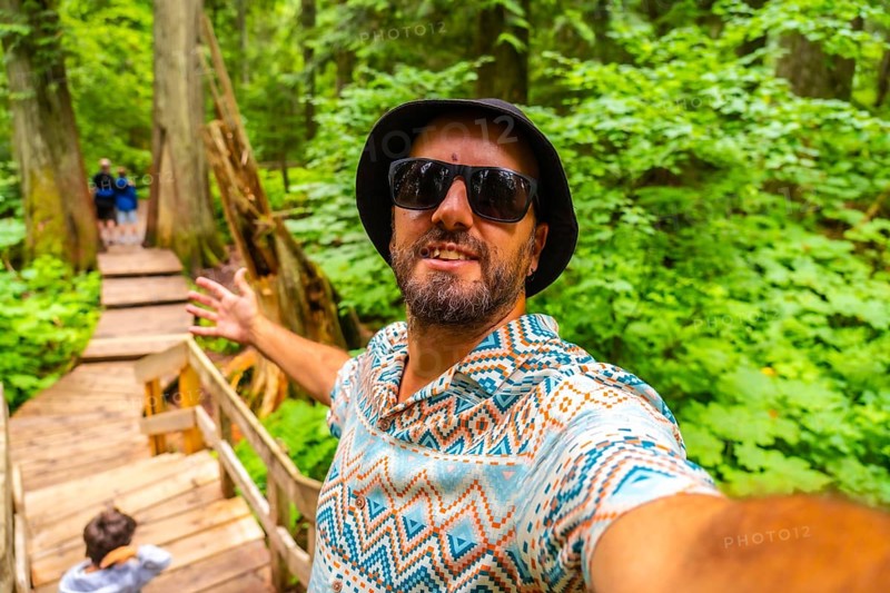 Happy tourist wearing sunglasses and a bucket hat taking a selfie while walking the giant cedars boardwalk trail in mount revelstoke national park, surrounded by lush greenery