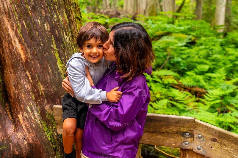 Mother kissing her smiling son while hiking the giant cedars boardwalk trail in mount revelstoke national park, british columbia, canada, surrounded by lush green ferns