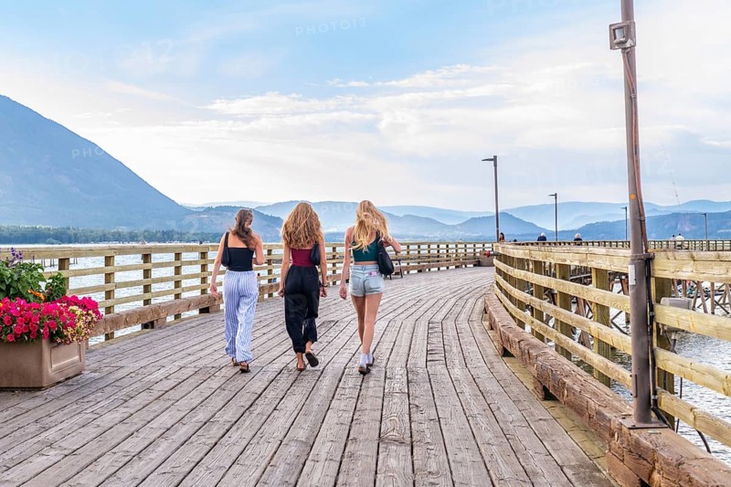 Three young women are enjoying a leisurely stroll along a scenic wooden pier in salmon arm, british columbia, surrounded by breathtaking views of the shuswap lake and surrounding mountains