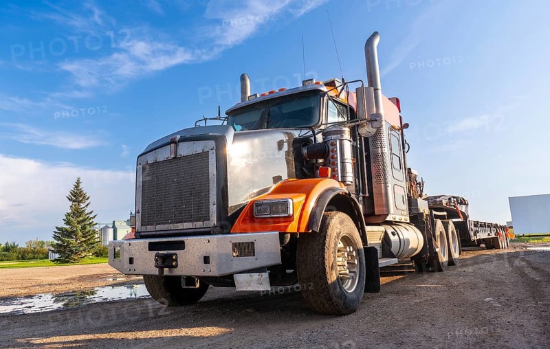 Gleaming orange and silver semi truck parked on a gravel road, bathed in the warm glow of late afternoon sunlight, against a backdrop of a clear blue sky and a serene rural landscape