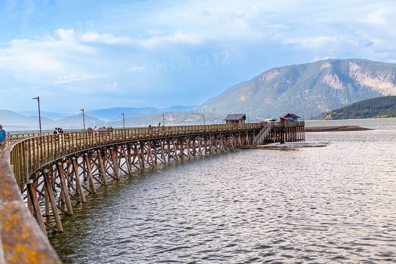 Tourists are enjoying a sunny day walking on the longest wooden wharf in north america, located in the city of salmon arm, british columbia, canada, with a mountain range in the background