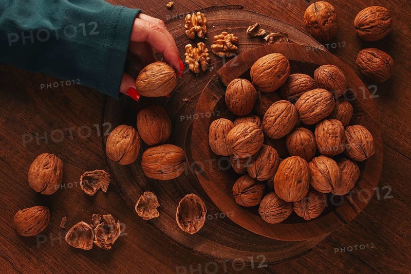 A woman's hand taking a walnut from a wooden bowl filled with walnuts. There are shells and walnuts scattered around