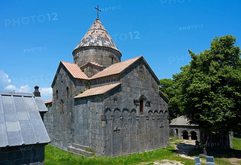 Medieval stone church with dome and cross under blue sky next to green trees, Sanahin monastery, Sanahin town, UNESCO World Heritage Site, Lorikeet province, Armenia