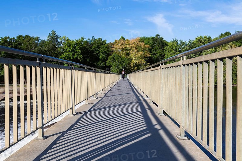 Electric scooter rider on pedestrian walkway on top of water flow control Moulin Neuf dam over Des Mille-Iles river in summer, Old Terrebonne, Quebec, Canada
