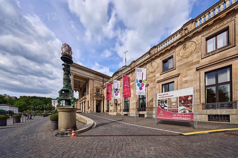 Spa hotel, access road, brick street, entrance area, flags on flagpoles, neoclassical style with elements of Art Nouveau, architect Friedrich von Thiersch, blue sky, cumulus clouds, Nimbostratus clouds, Kurhausplatz, Wiesbaden, state capital, district-free city, Hesse, Germany