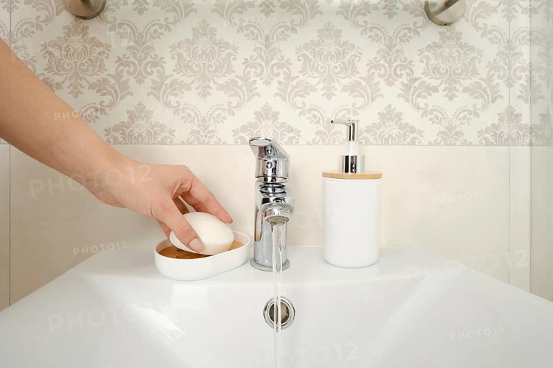 A hand reaches for a bar of soap placed on a dish near a sink. Water flows from a silver faucet into the basin. A soap dispenser sits beside the sink in a bathroom setting