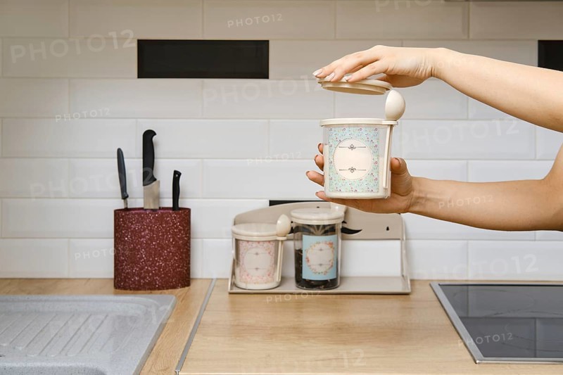 A person holds a container in one hand while opening the lid with the other. The kitchen has a clean counter with storage jars and knives visible nearby. The setting is bright and well-organized