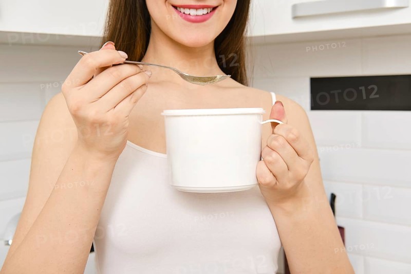 A woman stands in a kitchen holding large cup and spoon. She is smiling and appears to be preparing a hot drink. The kitchen has white tiles and basic utensils around her