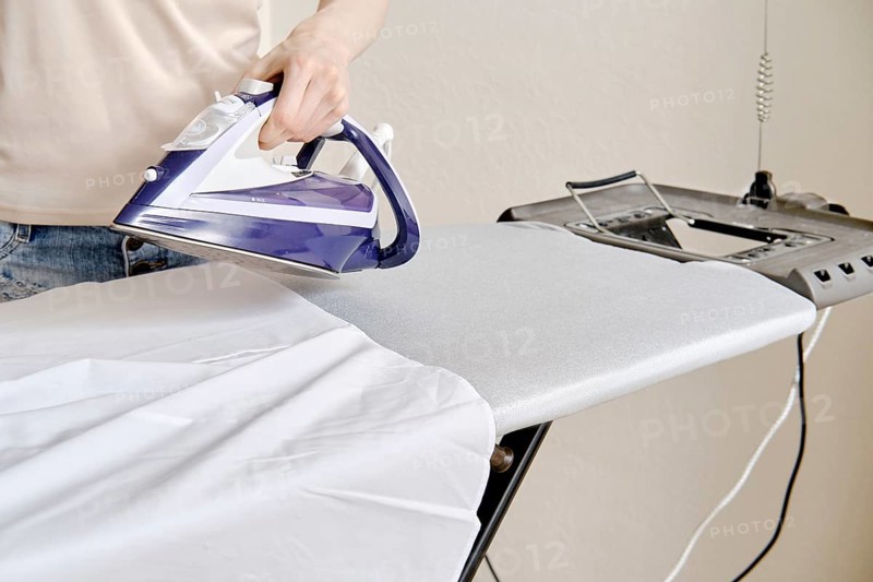 A person stands by an ironing board and uses an iron to press a shirt. The setting is a home environment during the afternoon. The task focuses on removing wrinkles from clothing