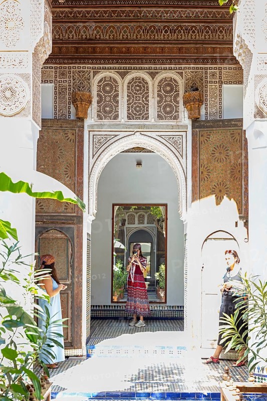 A traditional courtyard with arches, mirrors and decoration and two woman, Bahia Palace in Marrakech, Morocco