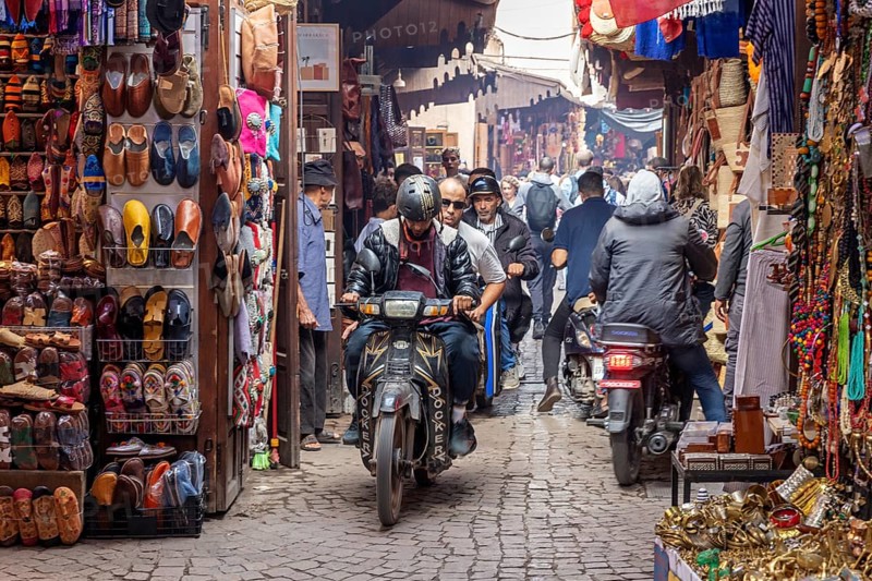 Bustling market with various stalls and motorcyclists navigating the alley