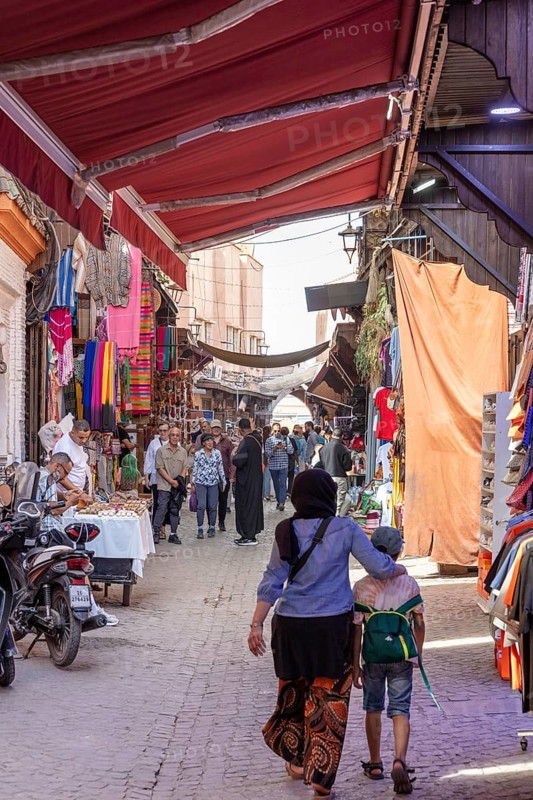Covered bazaar with colorful fabrics and people, a woman with a child walks through the market