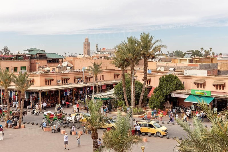 Bustling market scene with palm trees and buildings, a minaret and a blue sky in the background