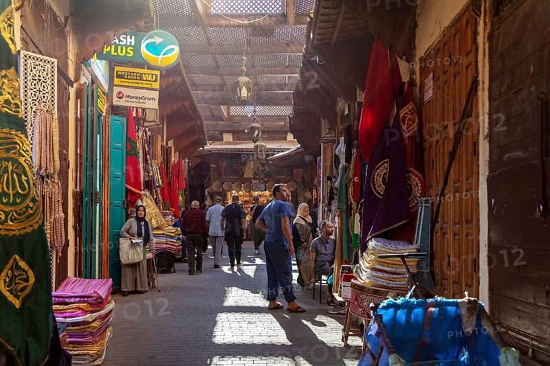Narrow market street with colorful textiles and carpets, passers-by stroll along