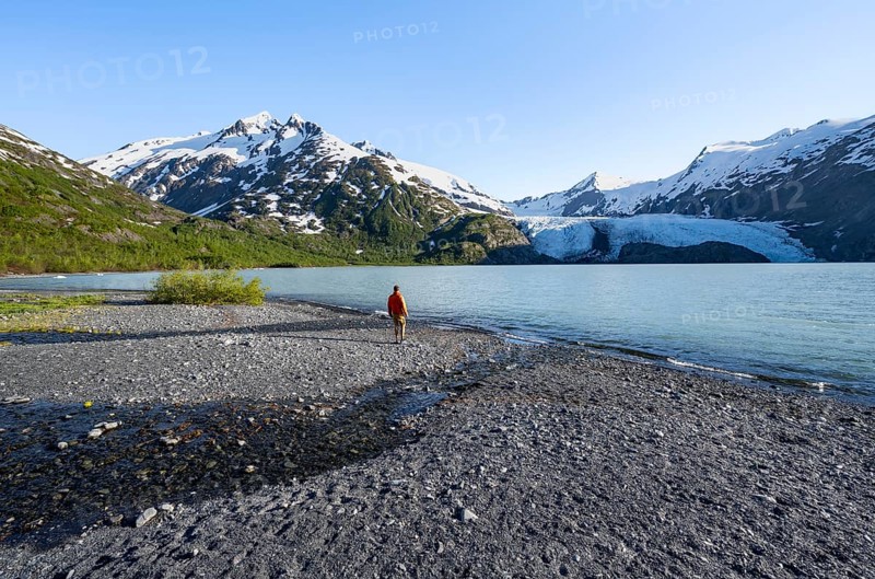 Young man on a pebble beach on a glacial lake, snowy mountains and Portage Glacier glacier on Portage Lake glacial lake, Chugach National Forest, Alaska, USA