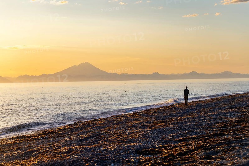Young man walking along the beach at sunset, view across Cook Inlet to white mountain peaks of Mount Redoubt, Aleutian Range Mountains, Anchor Point, Anchor River State Recreation Area, Alaska, USA