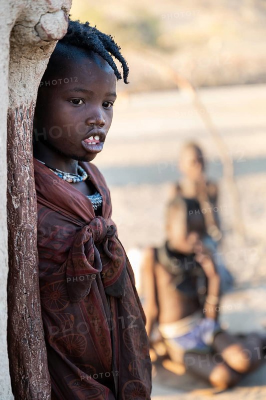 Himba girl leaning at a traditional clay hut, in the morning, traditional Himba village, Kaokoveld, Kunene, Namibia