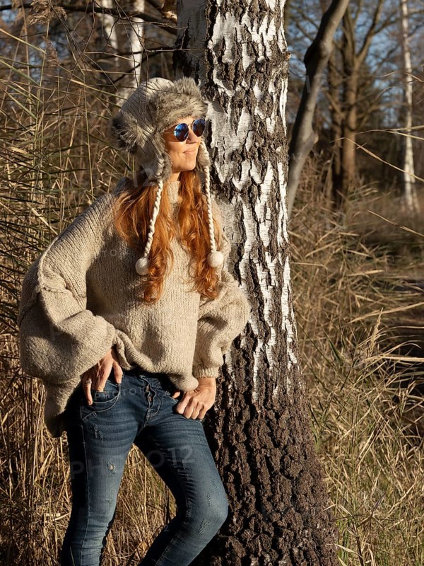 Slender pretty woman with fur hat and pompoms, red hair and warm winter colors between birch forest and reeds